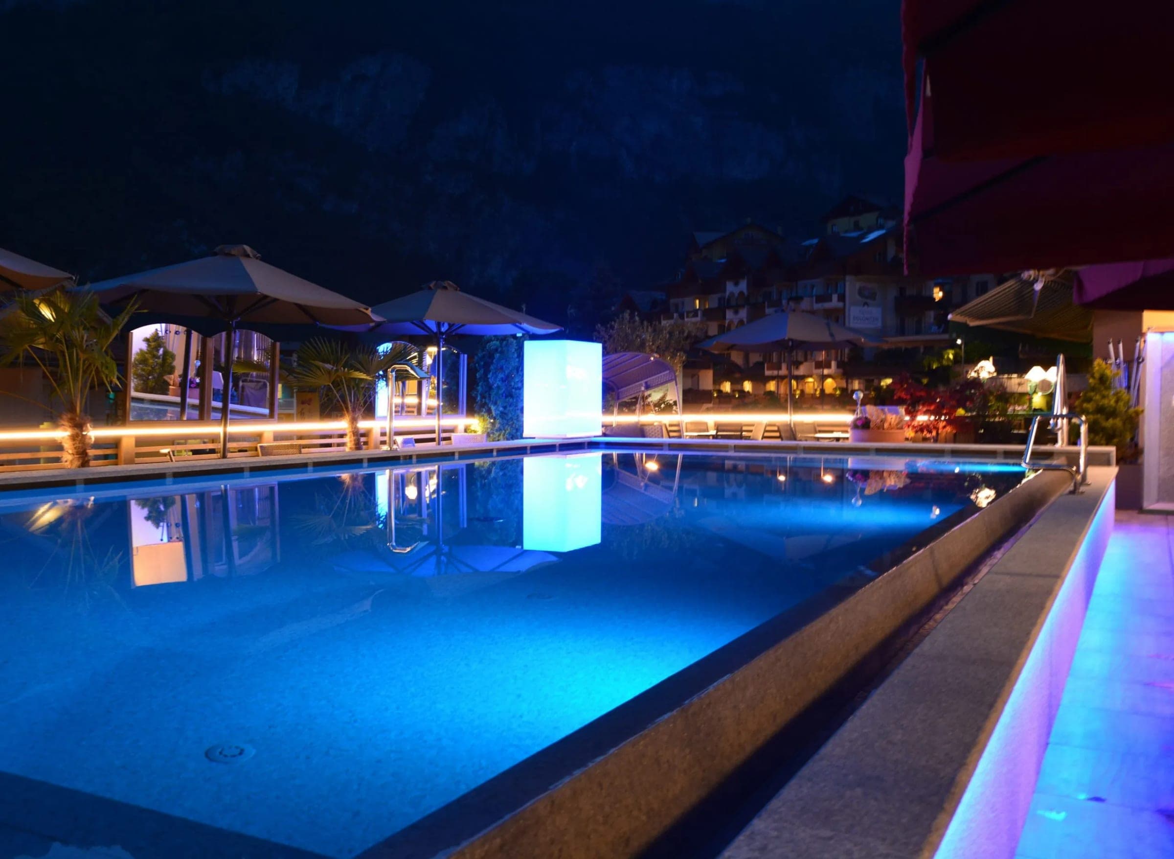 Zewnętrzny basen hotelu Alexander wieczorem, woda podświetlona na niebiesko, parasole i leżaki wokół