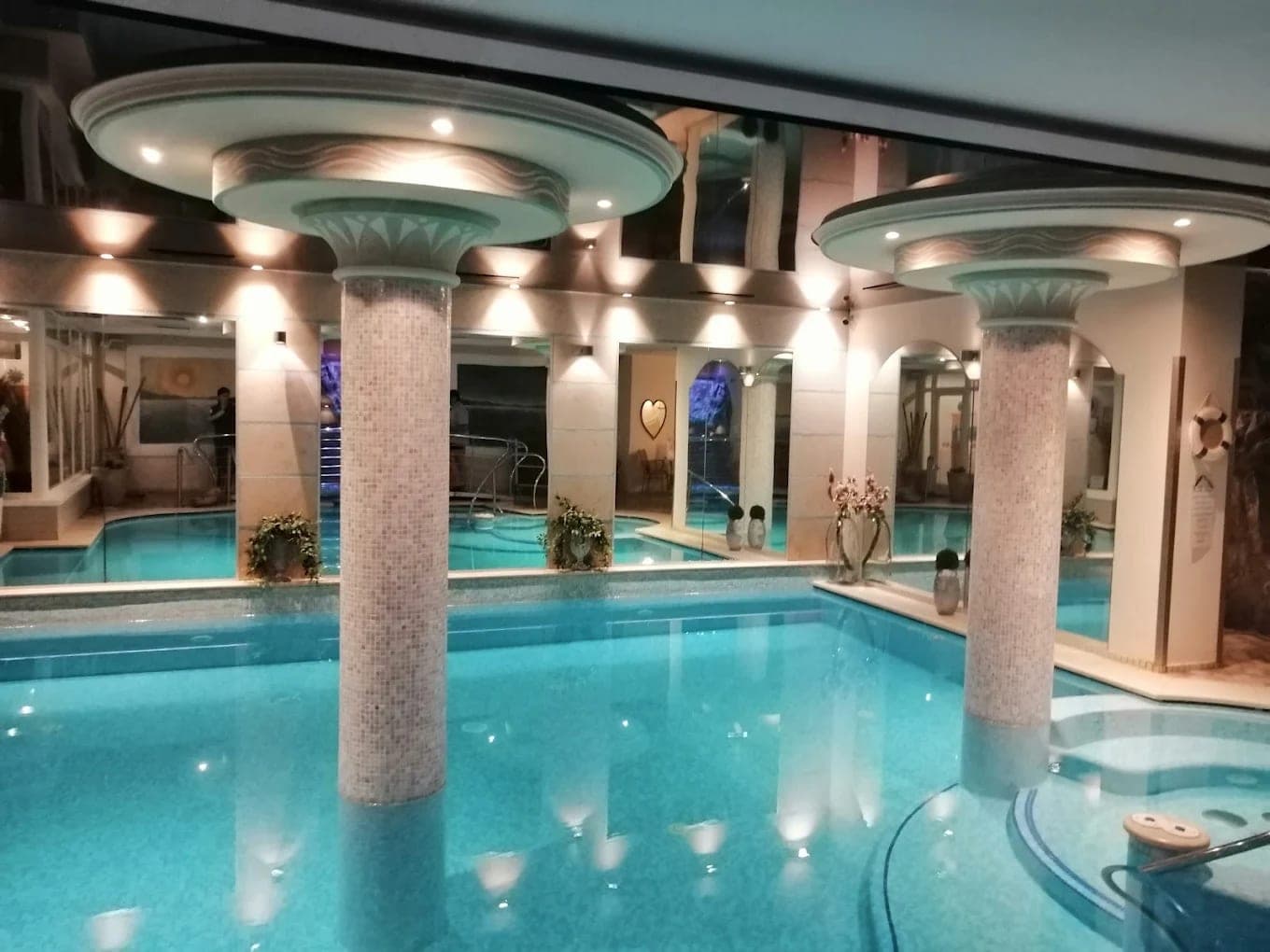 Kryty basen hotelu Alexander z mozaikowymi kolumnami, jacuzzi i podświetloną turkusową wodą
