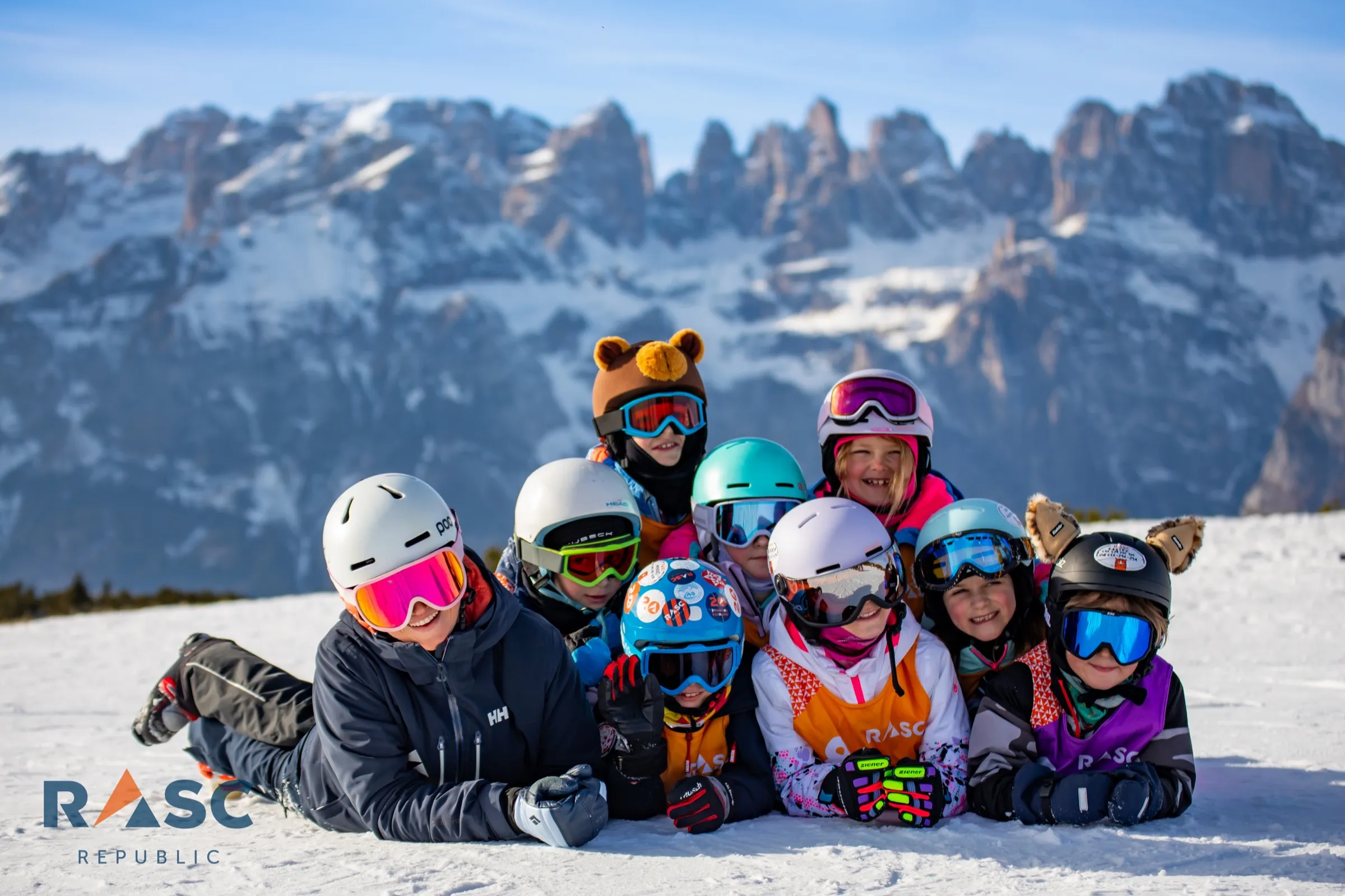 Grupa dzieci w kaskach narciarskich leżąca na śniegu, w tle turnie Dolomitów Brenty