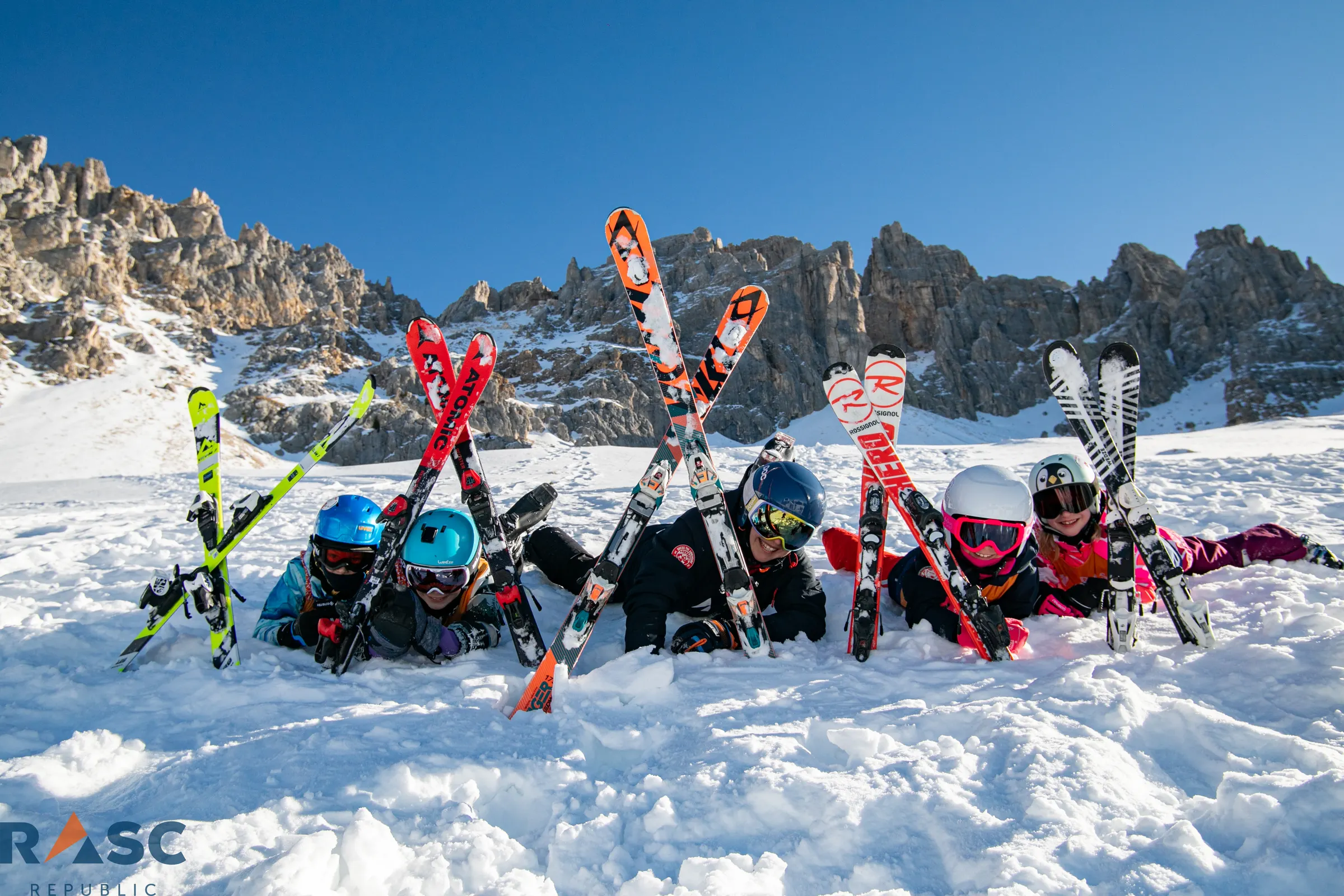 Grupa dzieci leży w śniegu z nartami uniesionymi w górę, za nimi skalna ściana Dolomitów