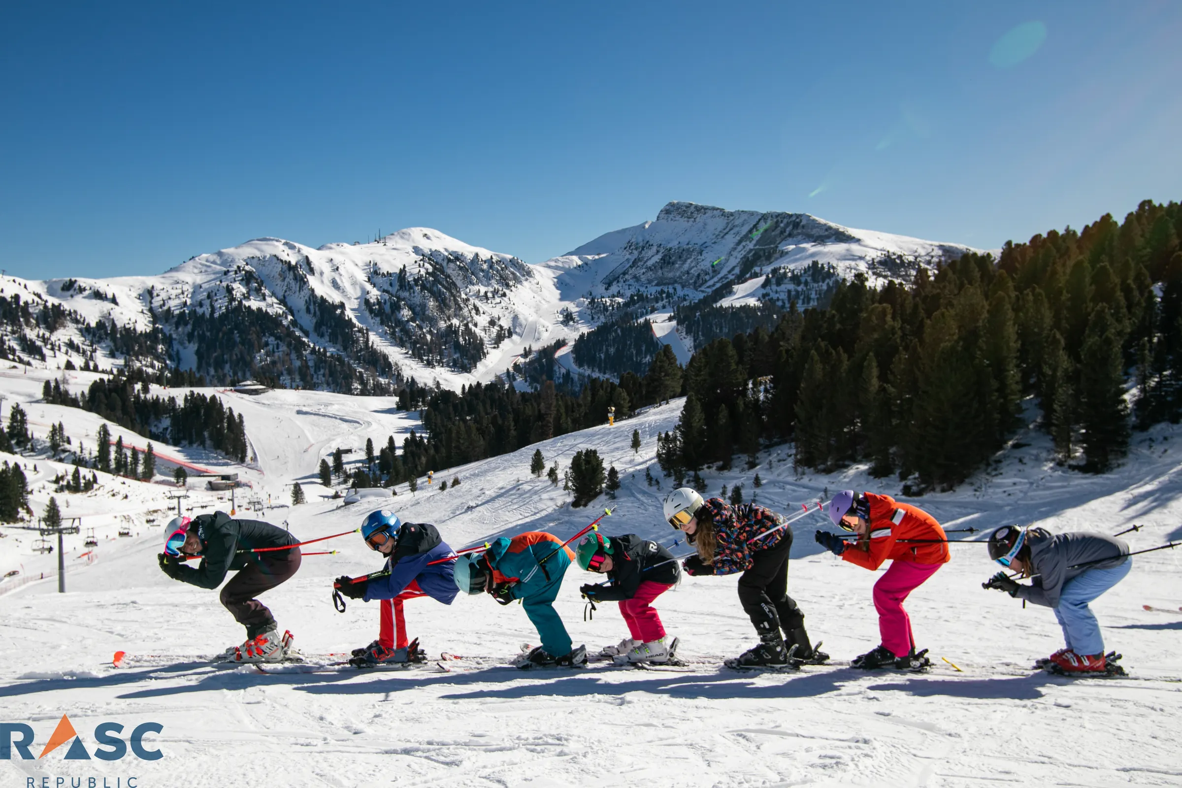 Sześcioro narciarzy w rzędzie w pozycji zjazdowej na stoku w Val di Fiemme, w tle panorama ośnieżonych szczytów