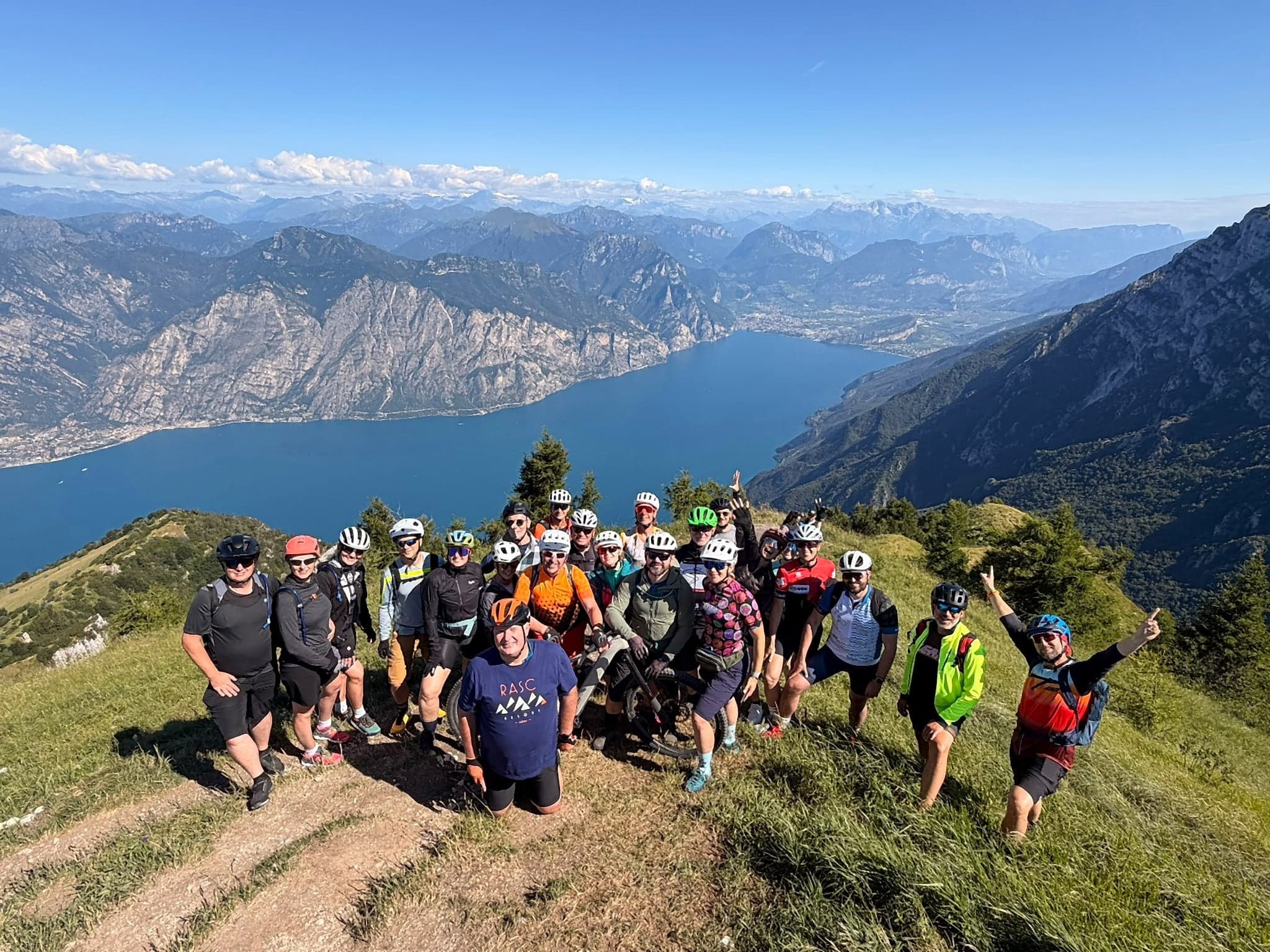 Grupa rowerzystów w kaskach pozuje z rowerami na trawiastym grzbiecie, w dole jezioro Garda i góry