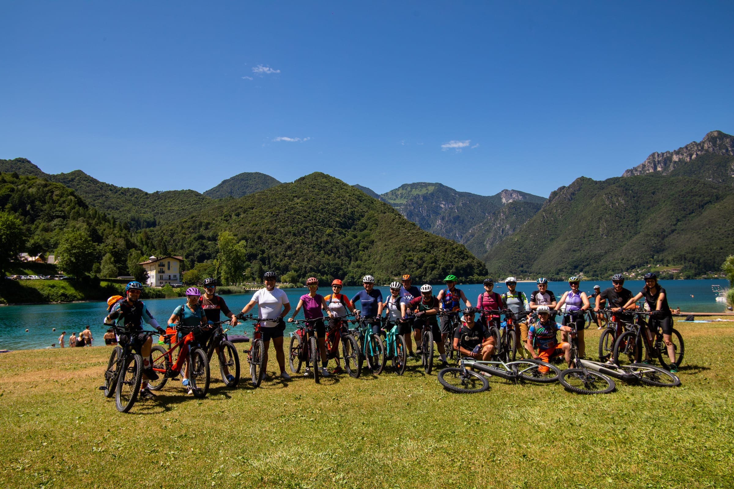 Grupa rowerzystów w kaskach stoi z rowerami na trawie nad turkusowym jeziorem Ledro, w tle zalesione góry
