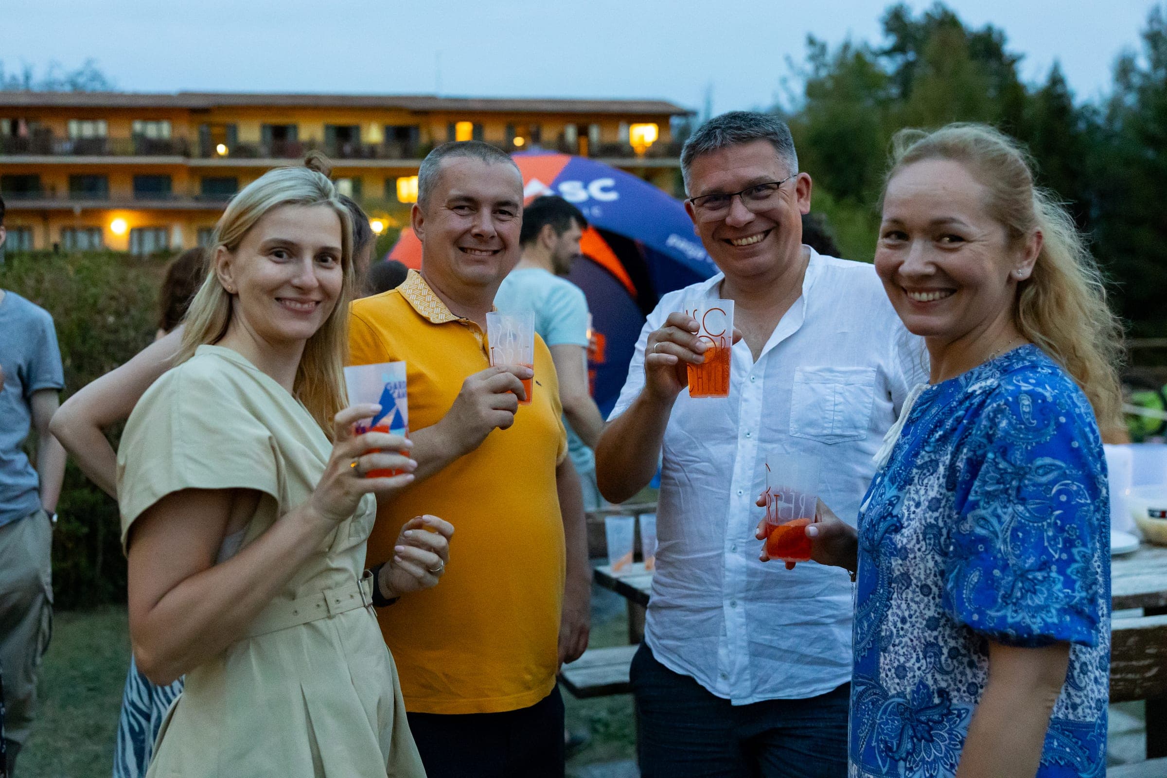 Czworo uśmiechniętych dorosłych wznosi toast pomarańczowymi drinkami w hotelowym ogrodzie o zmierzchu, za nimi namiot RASC i budynek hotelu