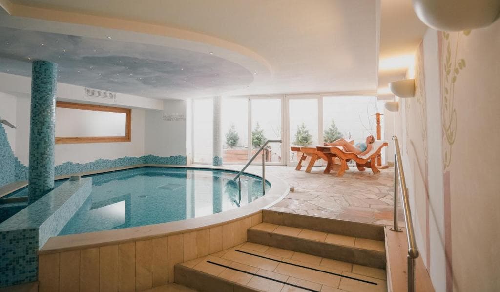 Kryty basen hotelu Olimpionico z mozaikową niecką, leżakami i dużymi oknami