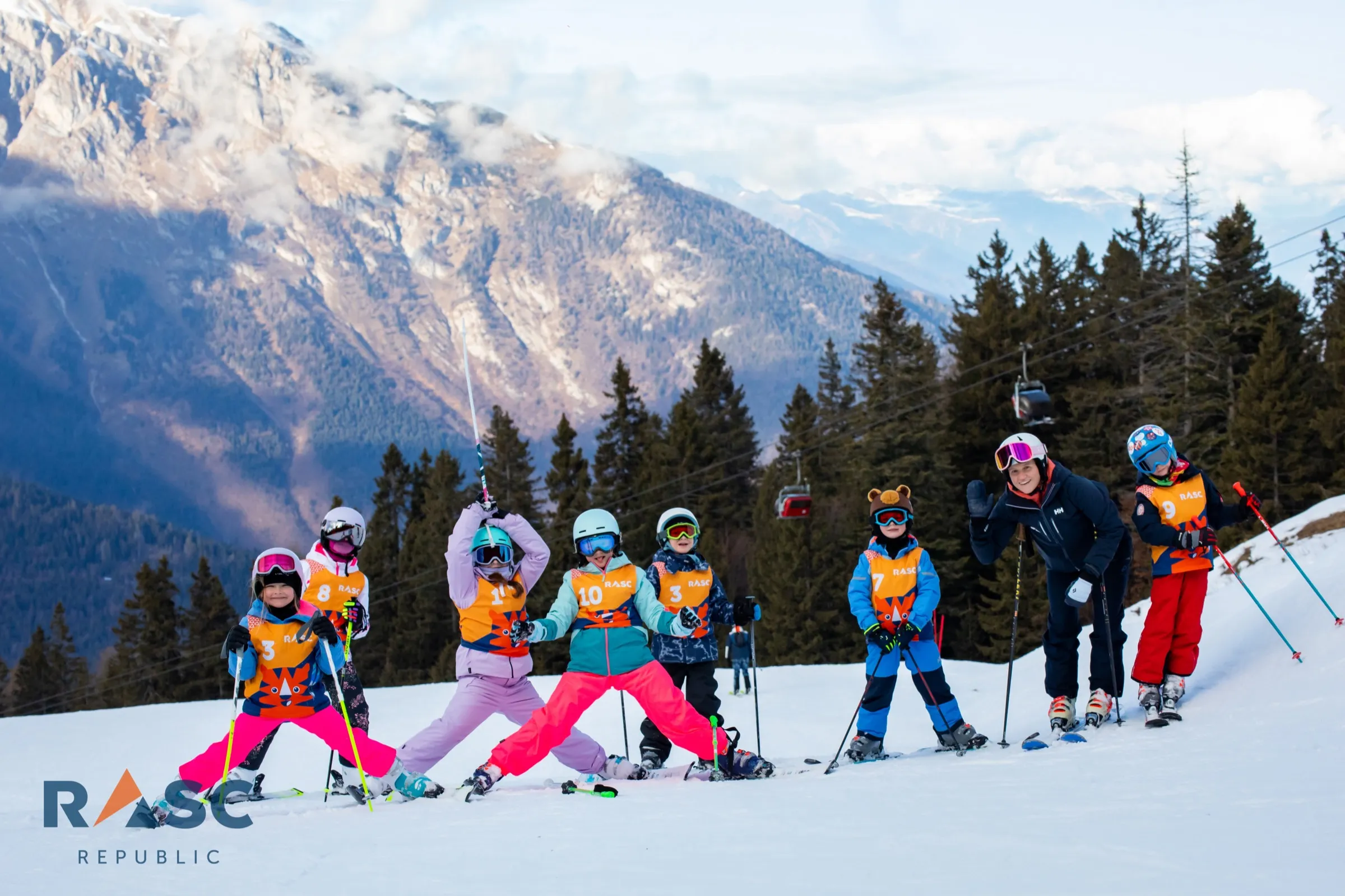 Grupa dzieci z instruktorem ustawiona w rzędzie na stoku, w tle zalesione zbocza i szczyty Dolomitów