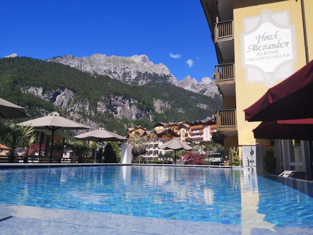 Hotel Alexander z szyldem na fasadzie i basenem zewnętrznym, w tle skalne granie Dolomitów Brenty
