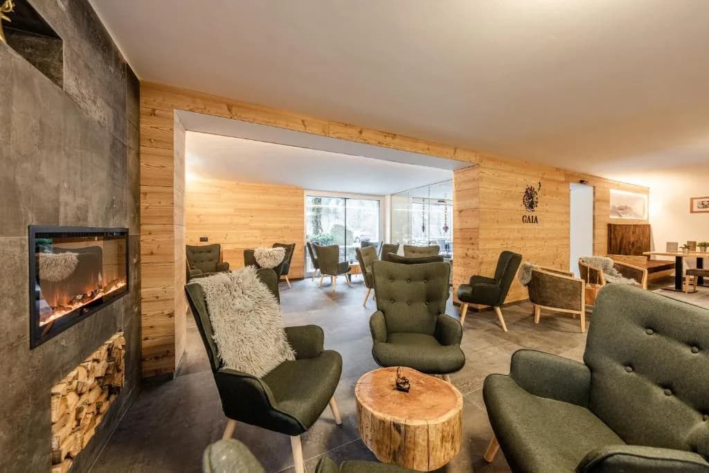 Lobby rezydencji Gaia z kominkiem, zielonymi fotelami i drewnianą zabudową ścian