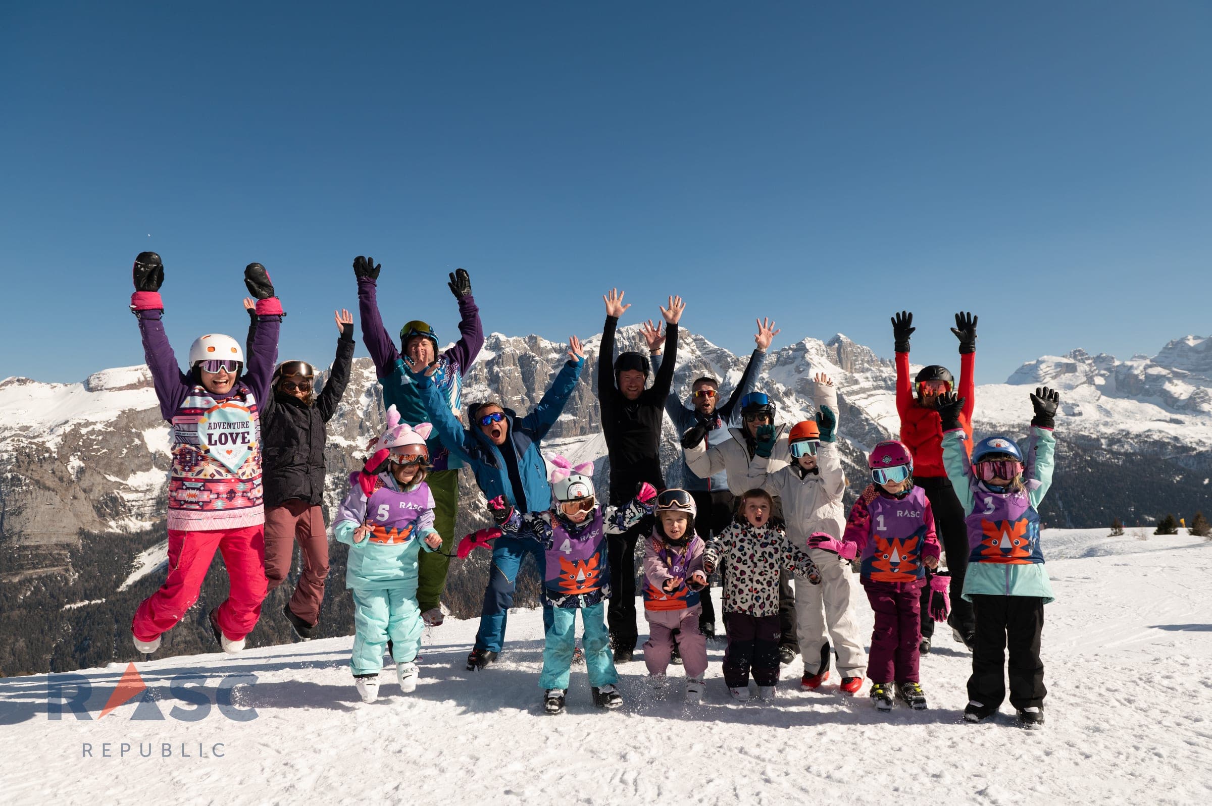 Cała grupa, dorośli i dzieci w kamizelkach TygrySKI, skacze z rękami w górze na ośnieżonym stoku, w tle panorama Dolomitów Brenty