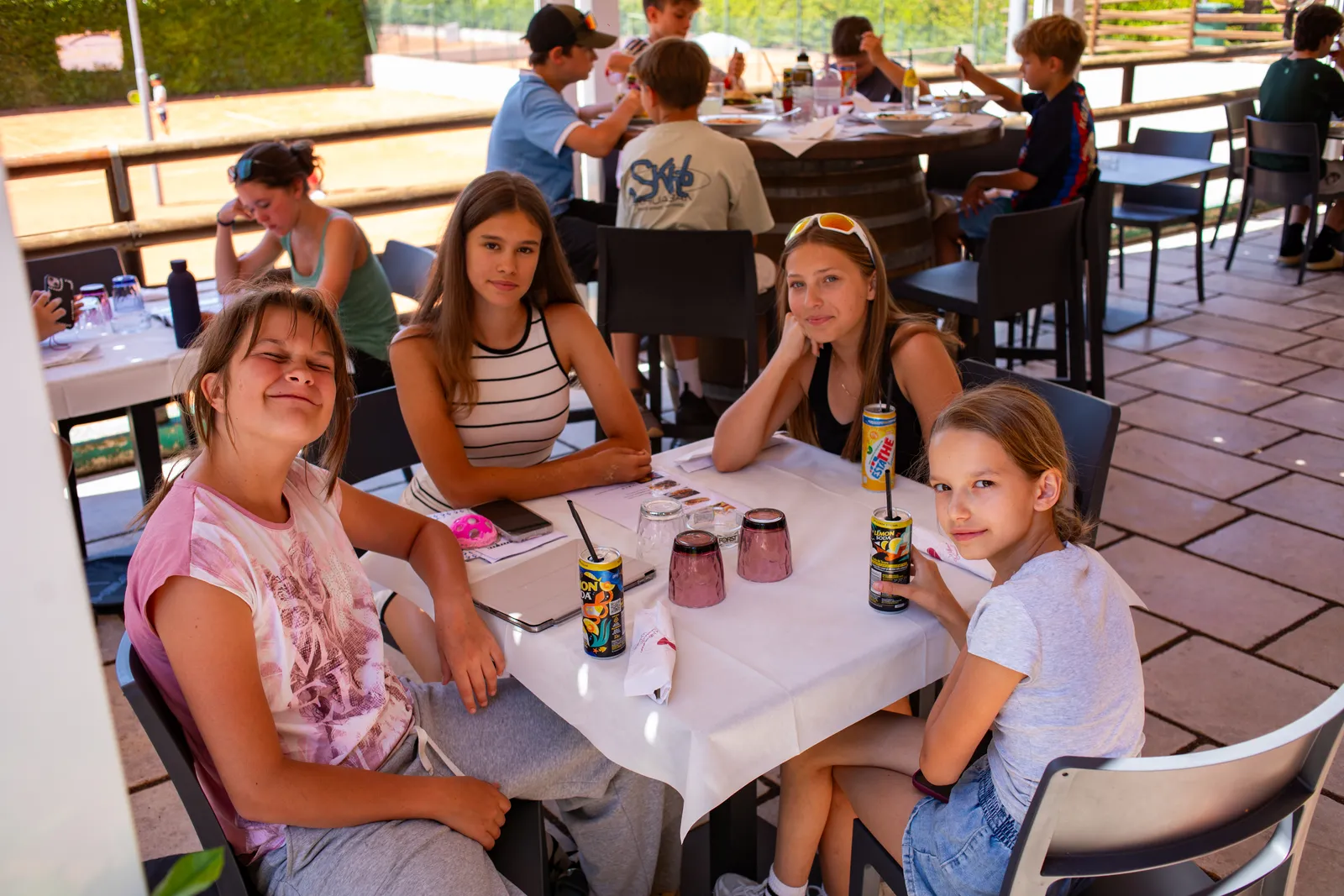 Cztery dziewczynki przy stoliku na zadaszonym tarasie restauracji czekają na lunch z napojami, za nimi inne dzieci jedzą przy okrągłym stole