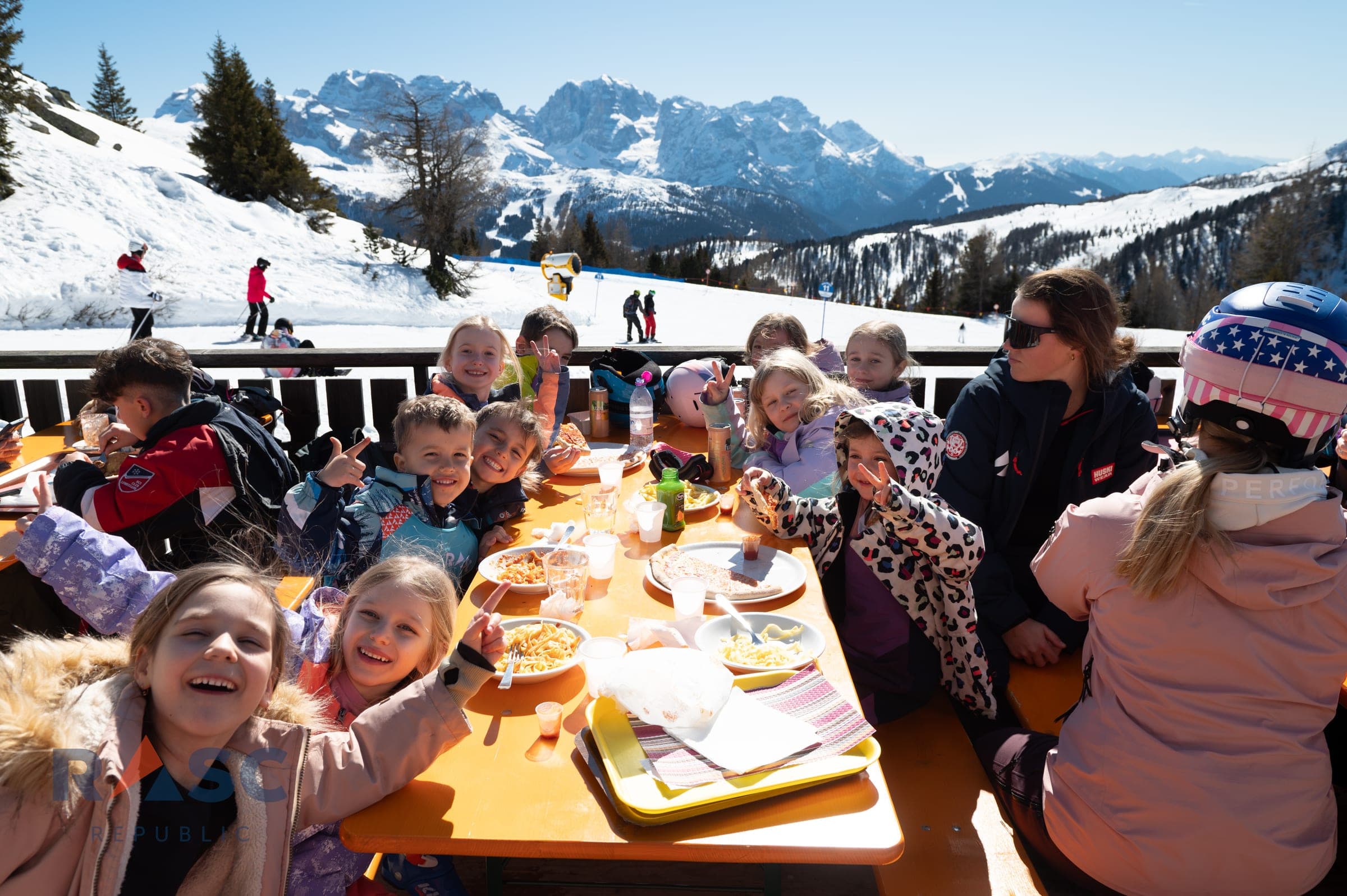 Dzieci z instruktorką przy stole na tarasie schroniska, na talerzach makaron i pizza, w tle panorama Dolomitów