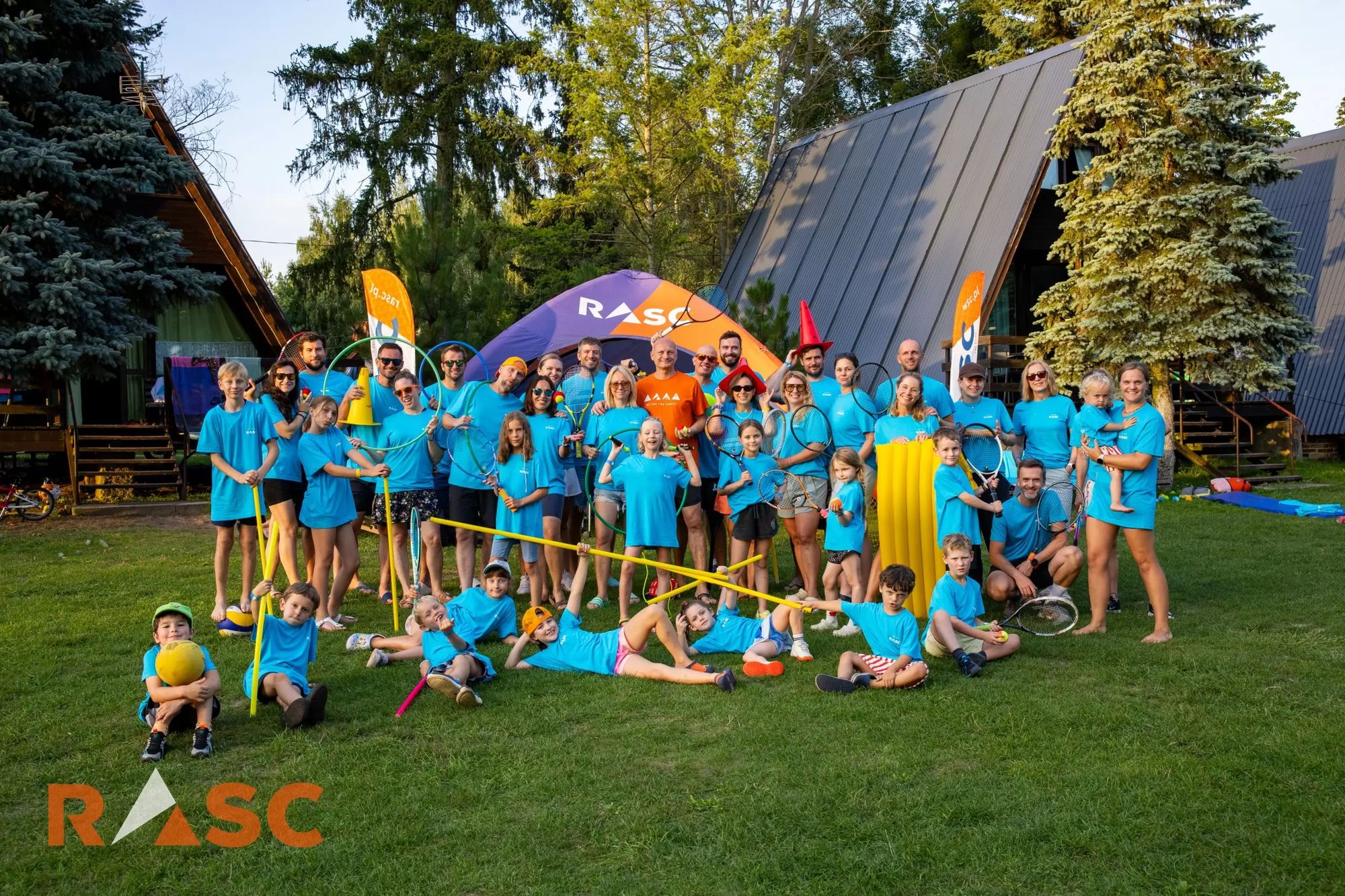 Uczestnicy Summer Family Camp w niebieskich koszulkach pozują z rakietami na trawie przed domkami ośrodka